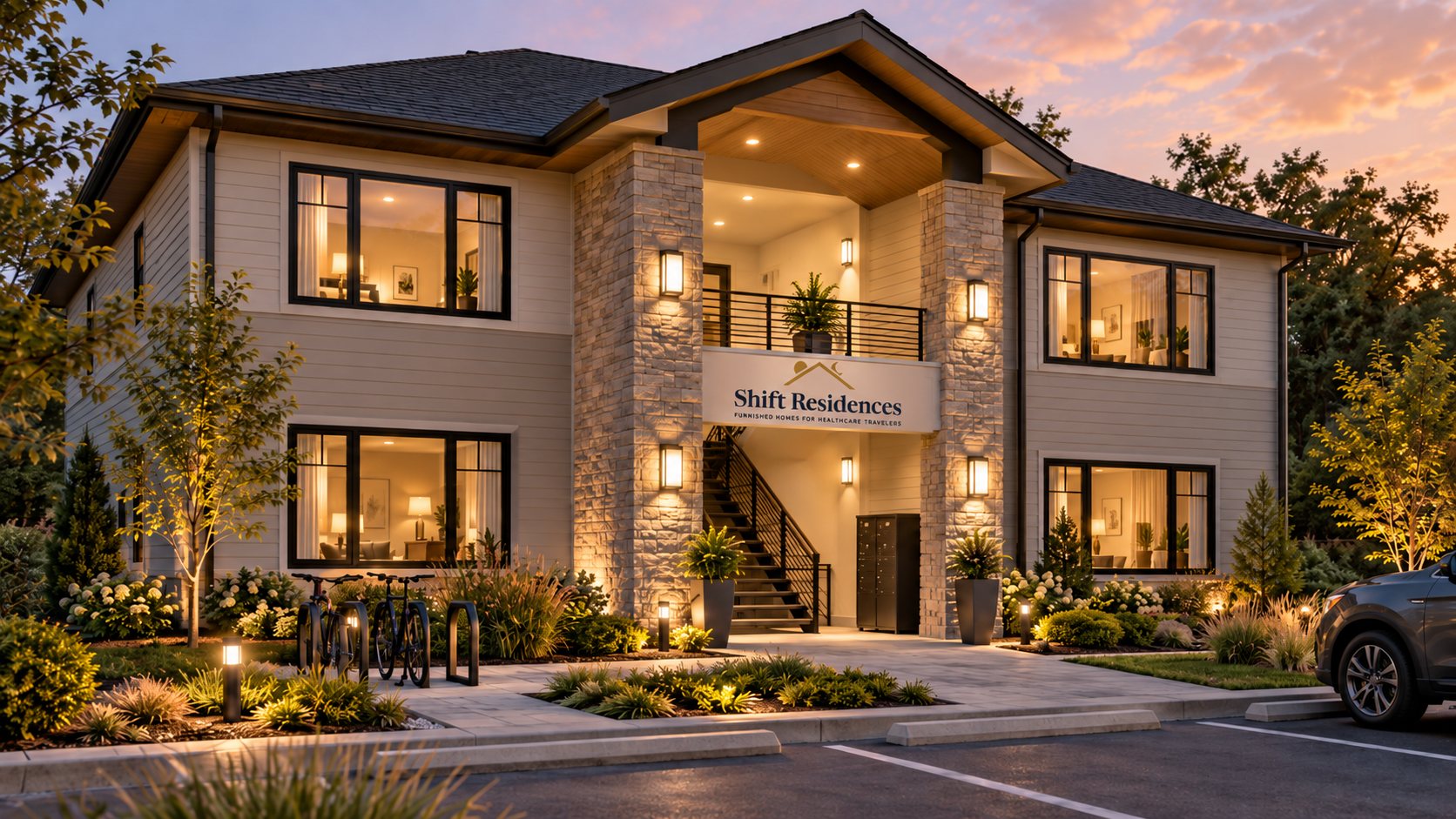 Concept rendering of a planned two-story Shift Residences building at dusk, with light siding, stone entry columns, a covered central stair, warm exterior lighting, landscaping, bike parking, mailboxes, and a parking area.